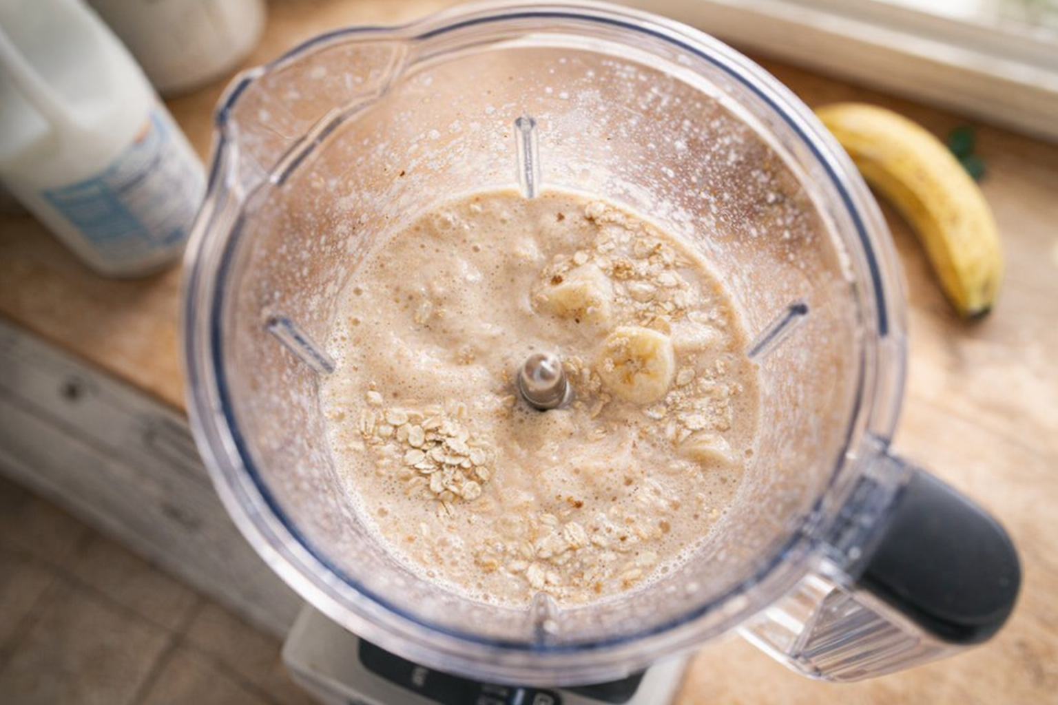 A blender with oats, banana, milk, and other ingredients being blended, overhead shot, bright kitchen light